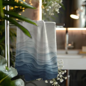 May include: A rectangular hand towel featuring a gradient pattern of wavy, flowing lines that transition from a light white at the top to a deep navy blue at the bottom. The towel is draped over a horizontal metal bar, displaying a smooth texture.
