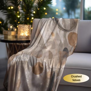 May include: A crushed velvet throw blanket featuring an abstract pattern of tan, beige, and dark gray circles, semi-circles, and thin vertical lines on a light cream background. The fabric has a soft, reflective sheen and a smooth texture.