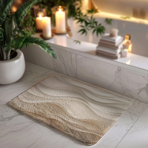 May include: A beige bath mat with a wavy pattern, resembling sand dunes. The mat is placed on a white tiled floor in a bathroom setting.