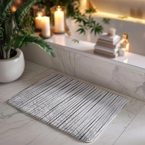 May include: A rectangular, silver bath mat with a ribbed design. The mat is placed on a marble tile floor. The background includes a potted plant, candles, and a bathtub.