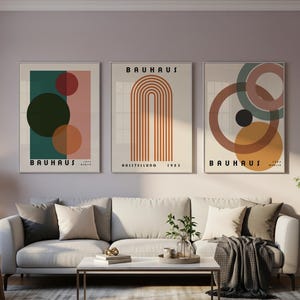 Può includere: Tre stampe d'arte da parete incorniciate con design Bauhaus. La stampa a sinistra mostra cerchi in verde, rosso e arancione su sfondo rosa. La stampa centrale mostra una serie di archi arancioni. La stampa a destra presenta cerchi sovrapposti in marrone, verde e rosa.