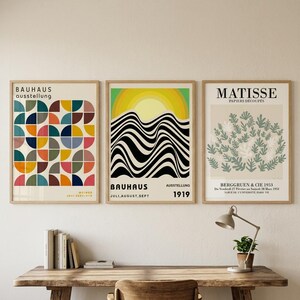 May include: Three framed art prints hanging on a wall. The left print features a colorful geometric pattern of semicircles. The center print displays a yellow sun over black and white wavy lines. The right print shows green Matisse-style foliage.