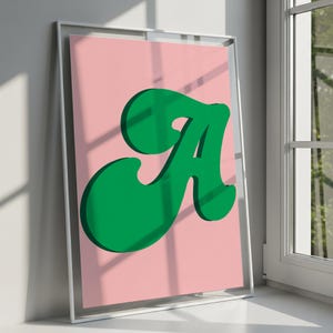 Könnte beinhalten: Ein gerahmter Kunstdruck mit einem großen, fetten, grünen Buchstaben A in einer stilisierten, abgerundeten Schriftart auf einem einfarbigen, hellrosa Hintergrund. Das Kunstwerk ist in einem dünnen weißen Rahmen gefasst und steht in einem hellen Raum.