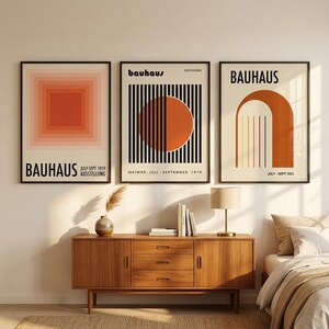 May include: Three framed Bauhaus exhibition posters displayed in a row. The left poster features concentric orange squares. The centre poster shows a large orange circle behind black vertical lines. The right poster displays an orange arch with lines.