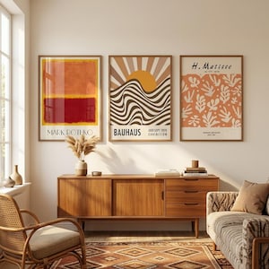 May include: Three framed art prints hanging on a wall above a wooden sideboard. The left print features orange and red colour blocks labelled Mark Rothko. The centre print shows a sun over wavy lines labelled Bauhaus. The right print displays orange leaf shapes labelled H. Matisse.