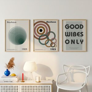 Może przedstawiać: Trzy oprawione grafiki w stylu Bauhaus. Grafiki przedstawiają geometryczne wzory w odcieniach zieleni, brązu i różu, z napisami "Bauhaus" i "1923". Trzeci nadruk głosi "GOOD VIBES ONLY". Grafiki są eksponowane na białej ścianie.