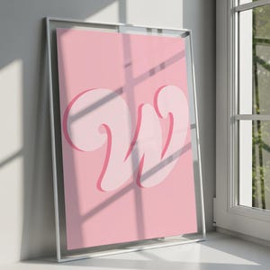 May include: A framed pink art print with a large, stylized letter "W" in a lighter shade of pink. The artwork is displayed in a silver frame, leaning against a wall near a window, with natural light casting shadows.