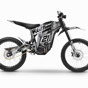 Può includere: Una moto da cross elettrica nera e grigia con un design grafico bianco e grigio. La moto ha una sella nera, un manubrio nero e ruote nere. La moto ha una catena nera e grigia e un motore nero e grigio. La moto ha un design grafico bianco e grigio con il testo "TALARIA" e "21" sul lato.