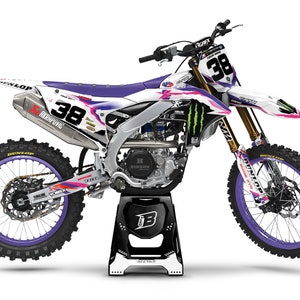 Peut inclure: Motocross blanche et violette avec un logo Monster Energy noir et vert sur le côté. La moto a une plaque d'immatriculation avec le numéro 38. La moto est sur un support noir et blanc.