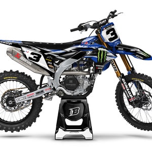 Può includere: Una moto da motocross nera e bianca con grafiche blu e bianche. La moto è su un cavalletto e ha un logo Monster Energy sul lato. La moto ha un pneumatico Dunlop sulle ruote anteriori e posteriori.
