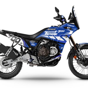 Può includere: Una moto Yamaha Ténéré 700 blu e bianca con una sella nera e uno scarico nero. La moto è dotata di un parabrezza grande e di un set di bauletti. La moto è decorata con le parole "Yamaha" e "Ténéré 700" in lettere bianche.