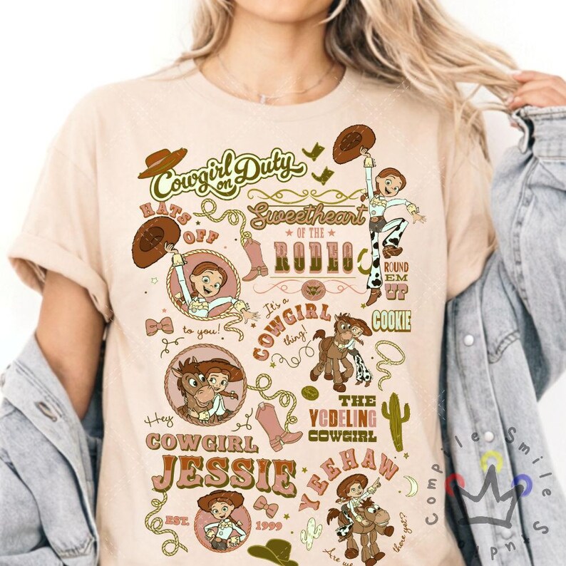 Op de afbeelding: Beige t-shirt met een kleurrijk grafisch ontwerp. Het ontwerp bevat de tekst "Cowgirl Duty" en "Sweetheart of the Rodeo", samen met illustraties van een cowgirl, laarzen, een paard en de naam "Jessie".