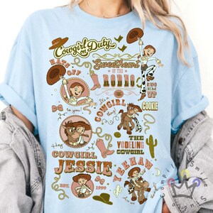 Op de afbeelding: Lichtblauw T-shirt met een grafisch ontwerp met de tekst "Cowgirl on Duty" en "Cowgirl Jessie". Het ontwerp bevat illustraties van een cowgirl, een paard, cowboylaarzen en een cowboyhoed. Het jaar 1999 is ook inbegrepen.