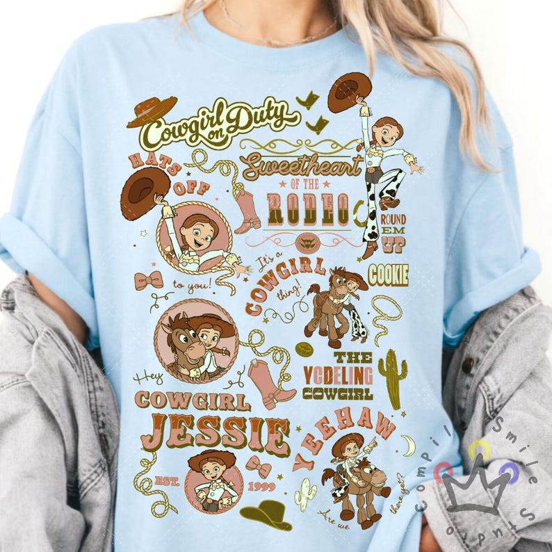 Op de afbeelding: Lichtblauw T-shirt met een grafisch ontwerp met de tekst "Cowgirl on Duty" en "Cowgirl Jessie". Het ontwerp bevat illustraties van een cowgirl, een paard, cowboylaarzen en een cowboyhoed. Het jaar 1999 is ook inbegrepen.
