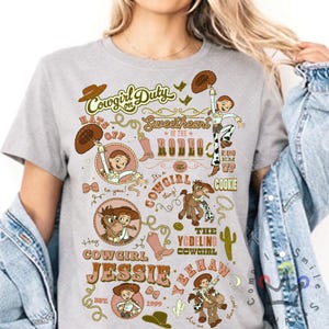 Op de afbeelding: Een lichtgrijs t-shirt met een grafisch ontwerp met de tekst "Cowgirl Duty" en "Sweetheart of the Rodeo". Het ontwerp bevat illustraties van een cowgirl, een paard en andere westerse elementen. Het shirt heeft ook de tekst "Hey Cowgirl Jessie" en "Yeehaw."