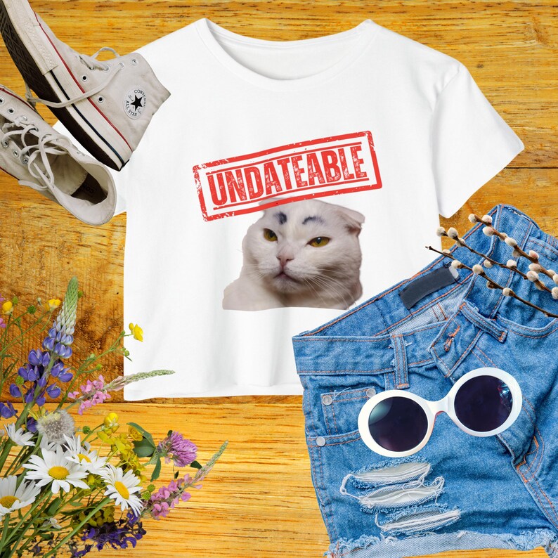 May include: White crop top with the word "UNDATEABLE" in a red stamp design, featuring a cat with drawn-on eyebrows. The image includes denim shorts, white sunglasses, and white sneakers. A bouquet of flowers is also present.
