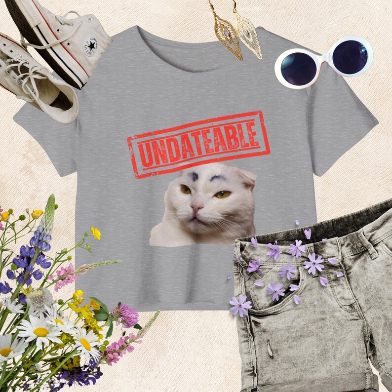 May include: A gray crop top with the word "UNDATEABLE" in a red stamp design, featuring a white cat with dark eyebrows. The image includes white sunglasses, gold earrings, denim shorts, and white sneakers.