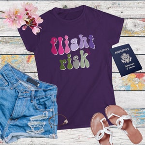 May include: A purple t-shirt with the words "flight risk" in a retro font. The shirt is paired with denim shorts, sandals, a passport, and a bracelet. Pink flowers are in the upper left corner.