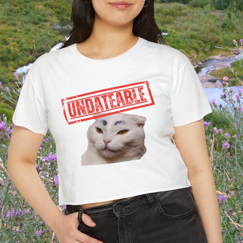 May include: White crop top t-shirt with the word "UNDATEABLE" in a red stamp design above a cat's face. The cat has white fur and yellow eyes. The background is a natural landscape.