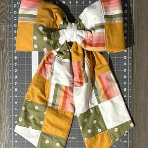 May include: A large, handmade bow tie featuring a patchwork design. The bow tie incorporates various colors, including mustard yellow, white, olive green, and pink stripes. Small white daisy patterns are scattered across the green patches. The bow tie is placed on a gray cutting mat.