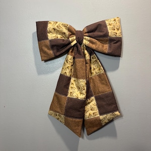Op de afbeelding: Een decoratieve stoffen strik met een patchworkontwerp van bruine en beige stoffen. Het patroon bevat vierkanten met een kleine stippenprint, een effen bruine paisley-textuur en een crèmekleurige achtergrond met een herhalende cowboylaarzenprint.