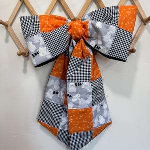 May include: A large decorative bow made of fabric featuring a patchwork design. The pattern consists of alternating squares of black and white gingham, orange fabric with small white dots, and a light gray fabric printed with white ghosts and the text BOO!