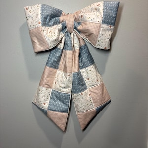 Op de afbeelding: Een grote decoratieve strik gemaakt van doorgestikte stoffen lappen. Het ontwerp bevat een herhalend patroon in stofblauw, zachtroze en crème, met bloemmotieven, kleine geometrische vormen en een subtiel ruitpatroon in de stof.