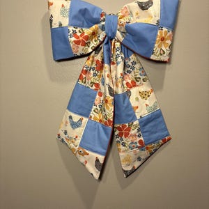 Könnte beinhalten: Eine große dekorative Schleife aus einem Patchwork aus Stoffquadraten. Das Design wechselt zwischen einfarbigen hellblauen Quadraten und einem cremefarbenen Stoff mit einem wiederkehrenden Muster aus bunten Blumen und verschiedenen Hühnern in Schwarz, Weiß und Braun.