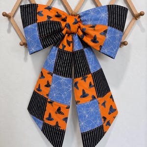 May include: A large fabric bow featuring a patchwork design of orange fabric with blue witch hat prints, light blue fabric with white spiderweb patterns, and black fabric with thin white vertical stripes, all stitched together in a grid pattern.
