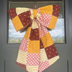 May include: A large, decorative bow made of patchwork fabric, featuring a mix of patterns and colors. The bow has a central knot and two large loops, with a combination of floral, dotted, and striped designs in shades of yellow, red, and white.