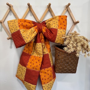 May include: A large fabric bow featuring a patchwork design of orange, yellow, and red squares. The patterns include small floral prints and a classic red and black gingham check. The bow is constructed from quilted fabric and hangs from a wooden rack.