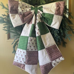 Op de afbeelding: Een grote, decoratieve strik gemaakt van gewatteerde stof, met een patchwork ontwerp in bruin, groen en wit. De strik is bevestigd aan een krans van groenblijvende takken, wat een feestelijke vakantiedecoratie creëert.