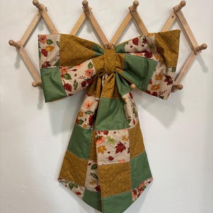 May include: A large decorative fabric bow featuring a patchwork design of autumn-themed patterns. The fabric includes squares with green gingham, solid mustard yellow with a crosshatch texture, and a cream background with colorful fall leaves.
