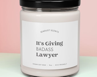 May include: A white candle jar with a black lid and a label that reads "Seadust Scents It's Giving Badass Lawyer Vegan Soy Wax - 9 oz - Eco Friendly"