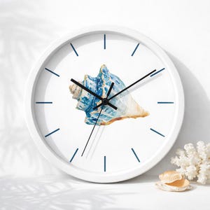May include: A round wall clock with a white frame and a white face featuring a central watercolor illustration of a blue and tan seashell. The clock has thin dark blue hour markers and three black hands indicating the time on the dial.