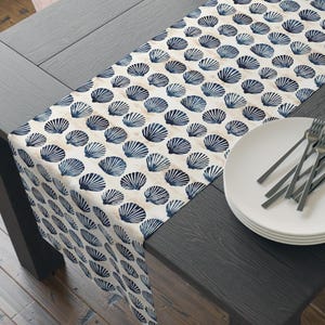 May include: A white table runner with a blue seashell pattern. The runner is laid on a dark wood table with a stack of white plates and silverware.
