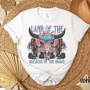 May include: White t-shirt with a graphic design featuring a bull skull with an American flag design, cacti, and the text "LAND OF THE FREE BECAUSE OF THE BRAVE" in red and blue.