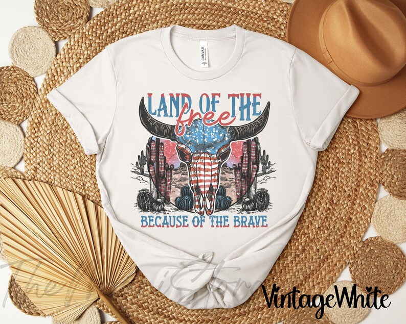 May include: A white t-shirt with a graphic design. The design features the text "LAND OF THE FREE" and "BECAUSE OF THE BRAVE", a bull skull with an American flag pattern, cacti, and a desert landscape. The shirt is tied at the waist.