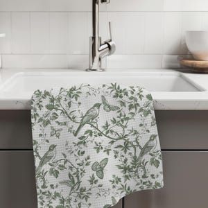 May include: A white kitchen towel with a green floral pattern featuring birds and butterflies. The towel is hanging on a kitchen sink.