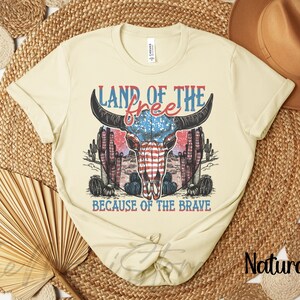 May include: A cream-coloured t-shirt with a graphic of a bull skull incorporating the American flag design. The text "LAND OF THE FREE" and "BECAUSE OF THE BRAVE" are in red and blue. The shirt has rolled-up sleeves.