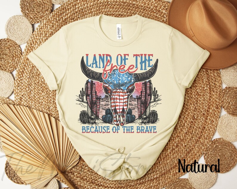 May include: A cream-coloured t-shirt with a graphic of a bull skull incorporating the American flag design. The text "LAND OF THE FREE" and "BECAUSE OF THE BRAVE" are in red and blue. The shirt has rolled-up sleeves.
