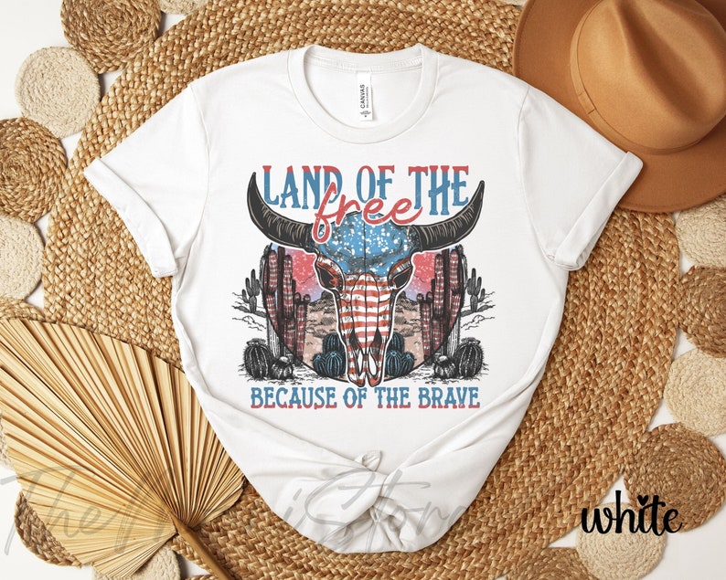 May include: White t-shirt with a graphic design featuring a bull skull with an American flag design, cacti, and the text "LAND OF THE FREE BECAUSE OF THE BRAVE" in red and blue.