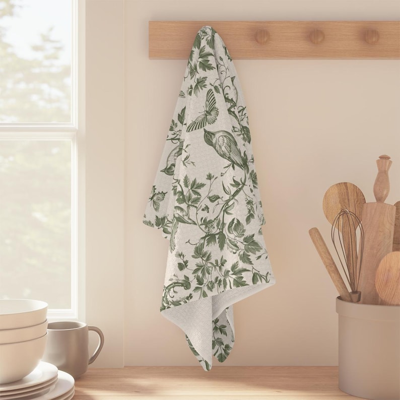 May include: A white kitchen towel with a green floral pattern featuring birds and butterflies. The towel is hanging on a wooden rack.
