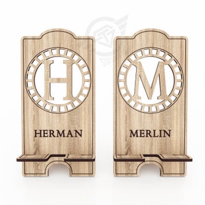 Op de afbeelding: Twee houten telefoonstandaards met een decoratief cirkelontwerp met de letters "H" en "M" in het midden. De standaards hebben de namen "HERMAN" en "MERLIN" gegraveerd onder de cirkels.