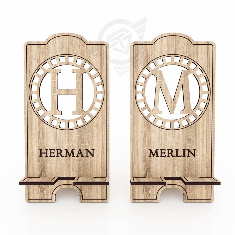 Op de afbeelding: Twee houten telefoonstandaards met een decoratief cirkelontwerp met de letters "H" en "M" in het midden. De standaards hebben de namen "HERMAN" en "MERLIN" gegraveerd onder de cirkels.