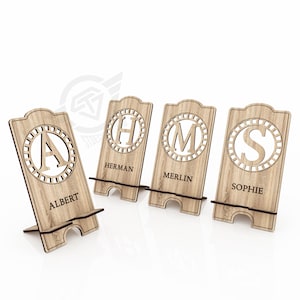 Op de afbeelding: Vier houten telefoonstandaards met gepersonaliseerde initialen en namen. De standaards zijn gemaakt van lichtbruin hout en hebben een cirkelvormig ontwerp met een decoratieve rand. De namen zijn "Albert", "Herman", "Merlin" en "Sophie".