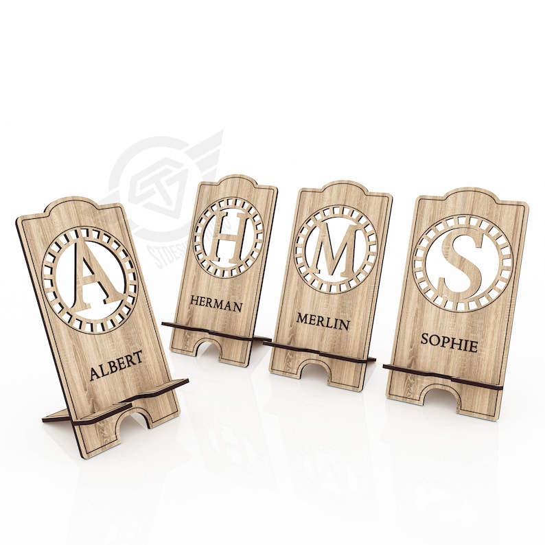 Op de afbeelding: Vier houten telefoonstandaards met gepersonaliseerde initialen en namen. De standaards zijn gemaakt van lichtbruin hout en hebben een cirkelvormig ontwerp met een decoratieve rand. De namen zijn "Albert", "Herman", "Merlin" en "Sophie".