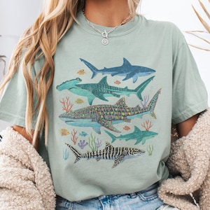 May include: A light green t-shirt featuring a watercolor illustration of various shark species, including hammerhead, whale, and tiger sharks. The design includes coral and small fish, creating an ocean-themed graphic.