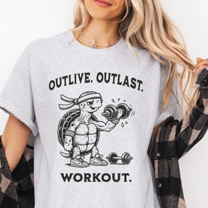 May include: Gray t-shirt with a black and white illustration of a turtle lifting weights. The text "OUTLIVE. OUTLAST. WORKOUT." is printed on the shirt.