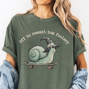 May include: Moss green t-shirt with a graphic of a snail wearing a witch hat on a skateboard. The text "Off to commit tom foolery" is arched above the snail. The shirt is a relaxed fit.